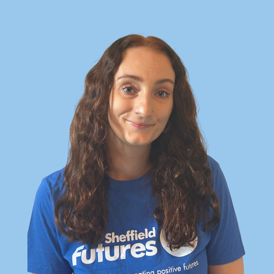 Kitty, a white woman with long brown hair, smiles at the camera. She is wearing a Sheffield Futures tshirt.