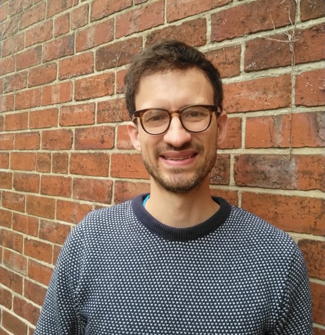 image shows Roo, a white person wearing round glasses and a grey jumper, smiling at the camera. They are against a brick wall backdrop.