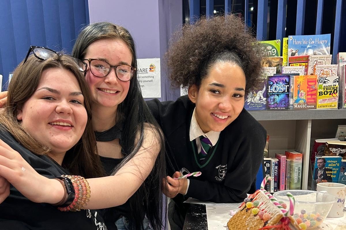 Three young people smile and hug each other, sat at a table in Wellbeing Cafe decorating gingerbread houses in the lead up to Christmas